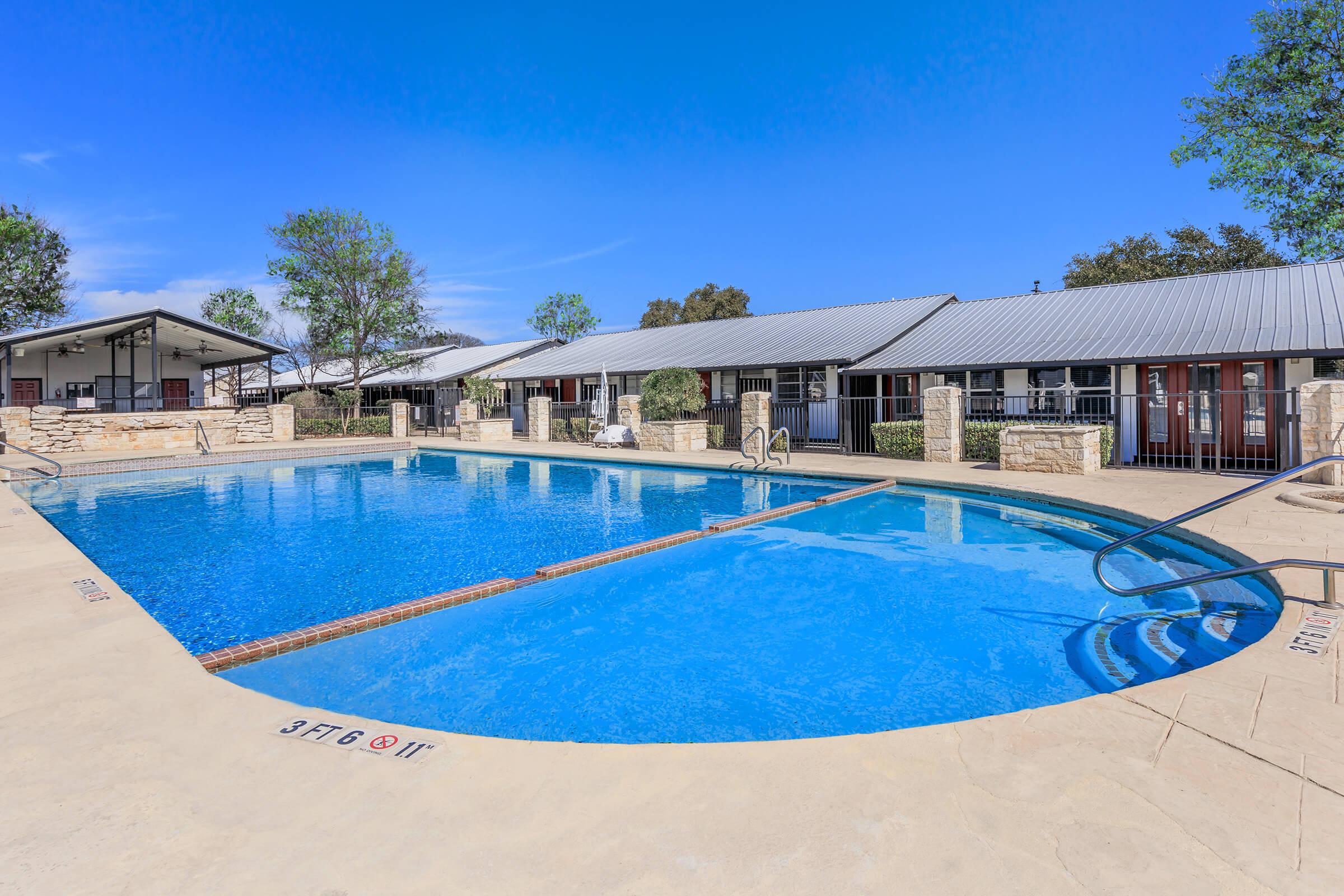 A clear blue swimming pool surrounded by a concrete deck, with steps leading into the water. Lush green trees are visible around the area. In the background, there are several buildings with metal roofs. The sky is bright and blue, indicating a sunny day.