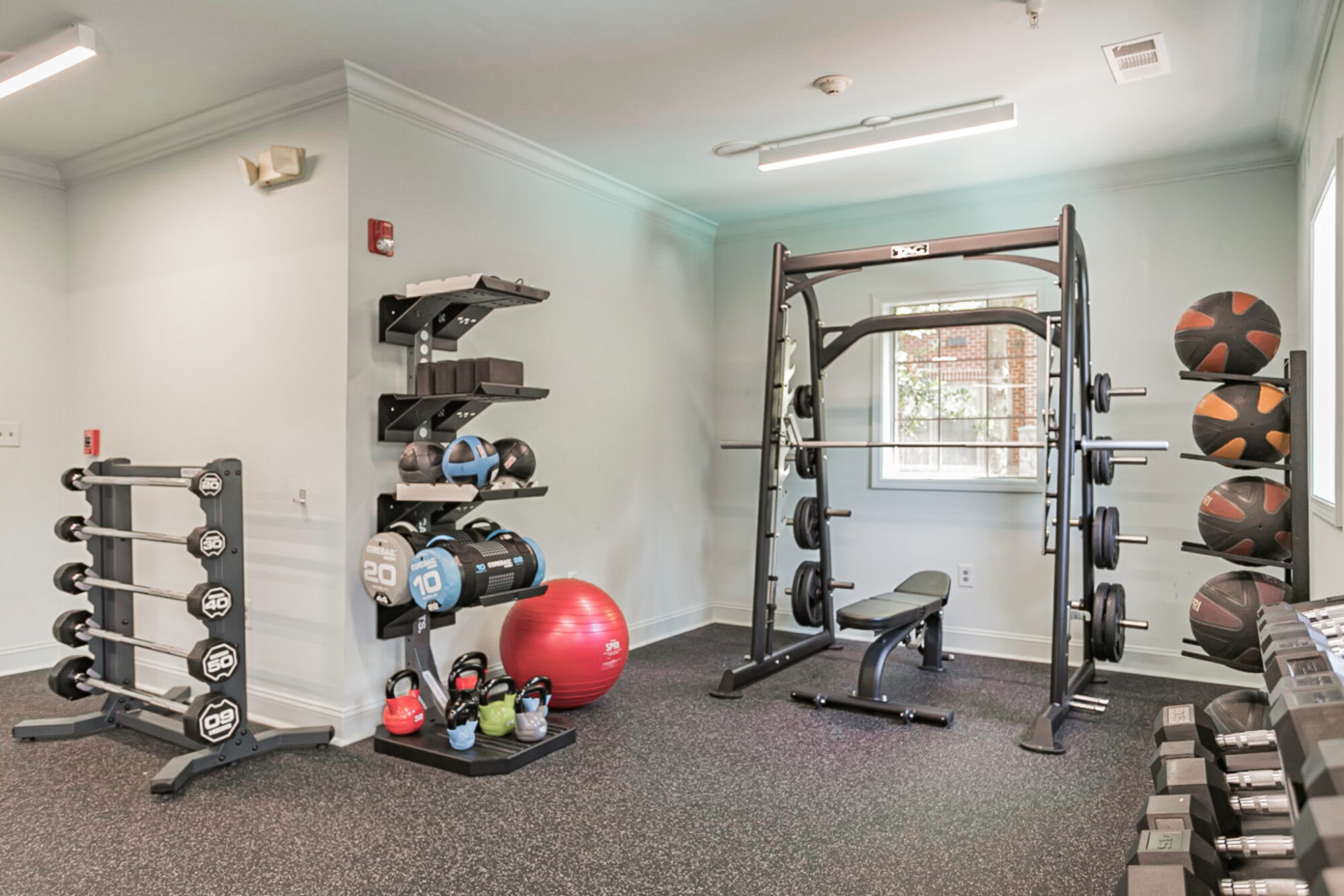 Small gym with free weights, kettlebells, medicine balls, a red exercise ball, a squat rack with weights, and dumbbells in front of a window. The room has light walls and black rubber flooring.