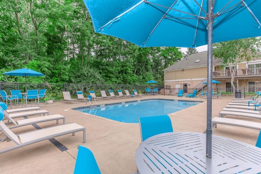A swimming pool area surrounded by lounge chairs and blue umbrellas, nestled in a lush green environment. The pool is clear and inviting, with a few chairs placed around the perimeter for relaxation. A table with matching blue chairs is in the foreground, offering a spot for guests to gather.