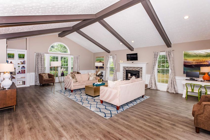 A spacious, modern living room featuring large windows, high vaulted ceilings with wooden beams, stylish sofas, and a coffee table. There is a cozy fireplace against one wall, and decorative elements include artwork and plants. The floor is a light wood, and the overall color scheme is neutral with pops of color.