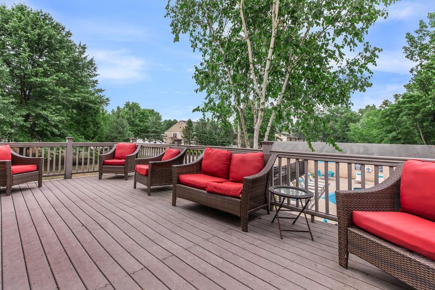 A spacious wooden deck with several wicker chairs featuring red cushions, surrounded by green trees and a clear blue sky.