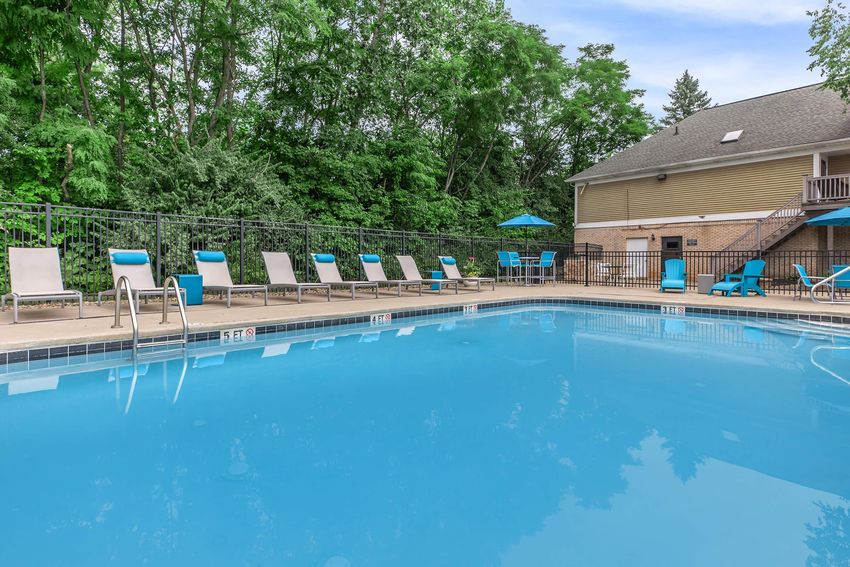 A clear blue swimming pool surrounded by lounge chairs, with a backdrop of lush greenery and a building in the distance.