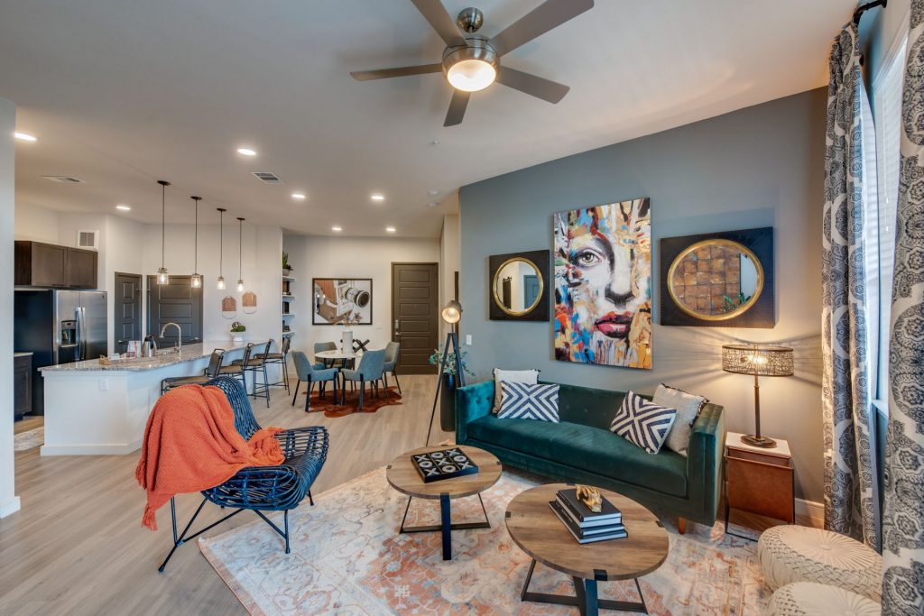 Living room area with kitchen access, wood style flooring, couch and chair seating, and wall paintings