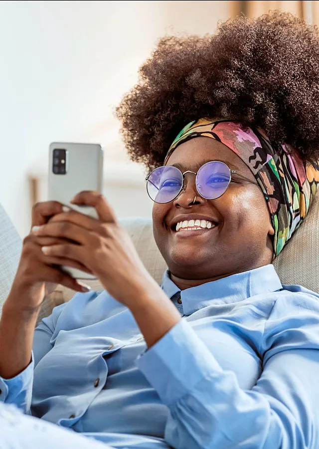 ReNew Minnehaha happy woman relaxing on a cozy sofa, browsing a smartphone with sunlight streaming in, exuding comfort and leisure.