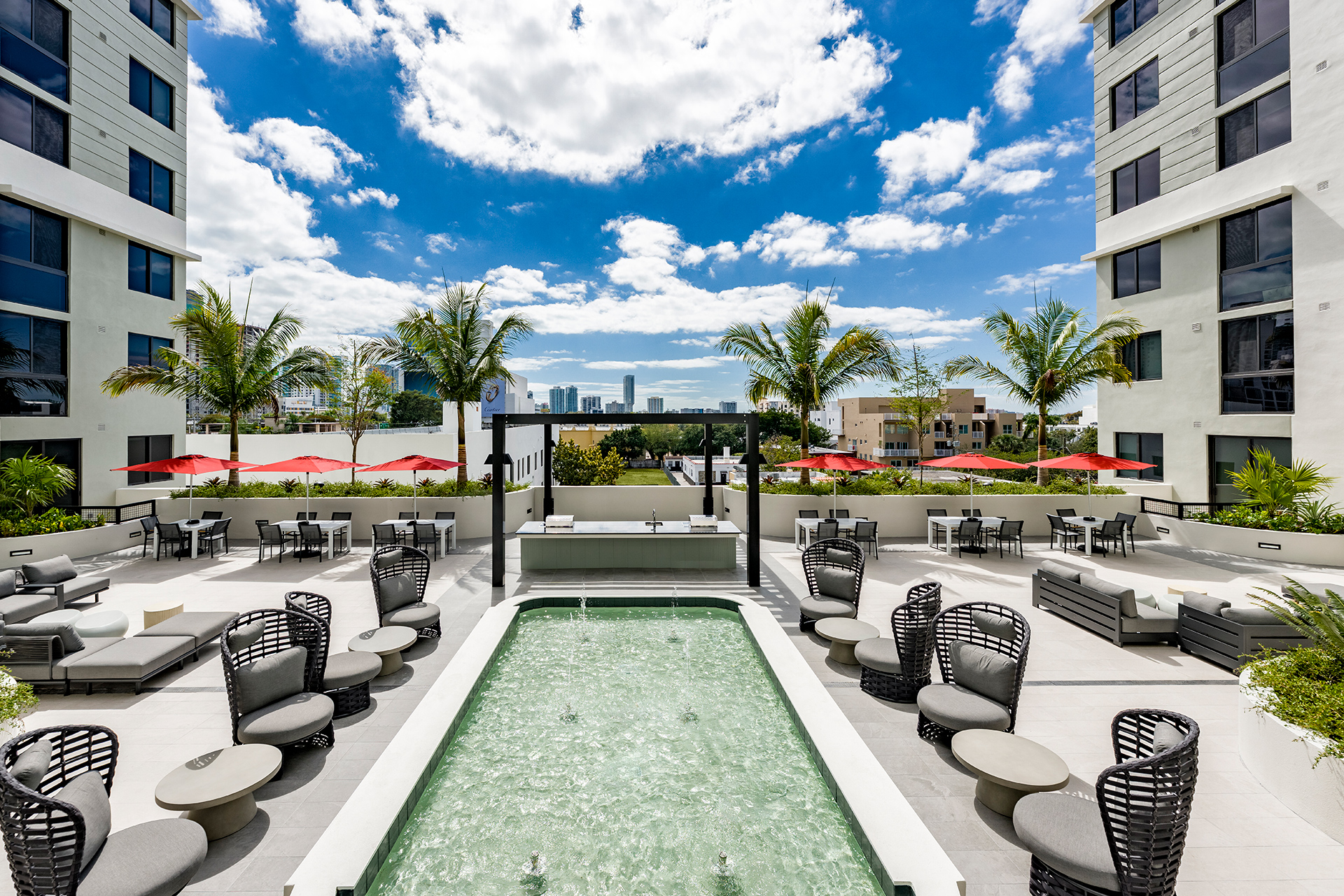 Two-Bedroom Apartments in Little Havana, FL - Gallery Marti Park - Rooftop Courtyard With Water Feature, Lounge Chairs, Red Umbrellas, Palm Trees, and Open Skyline Views Between Modern Buildings.