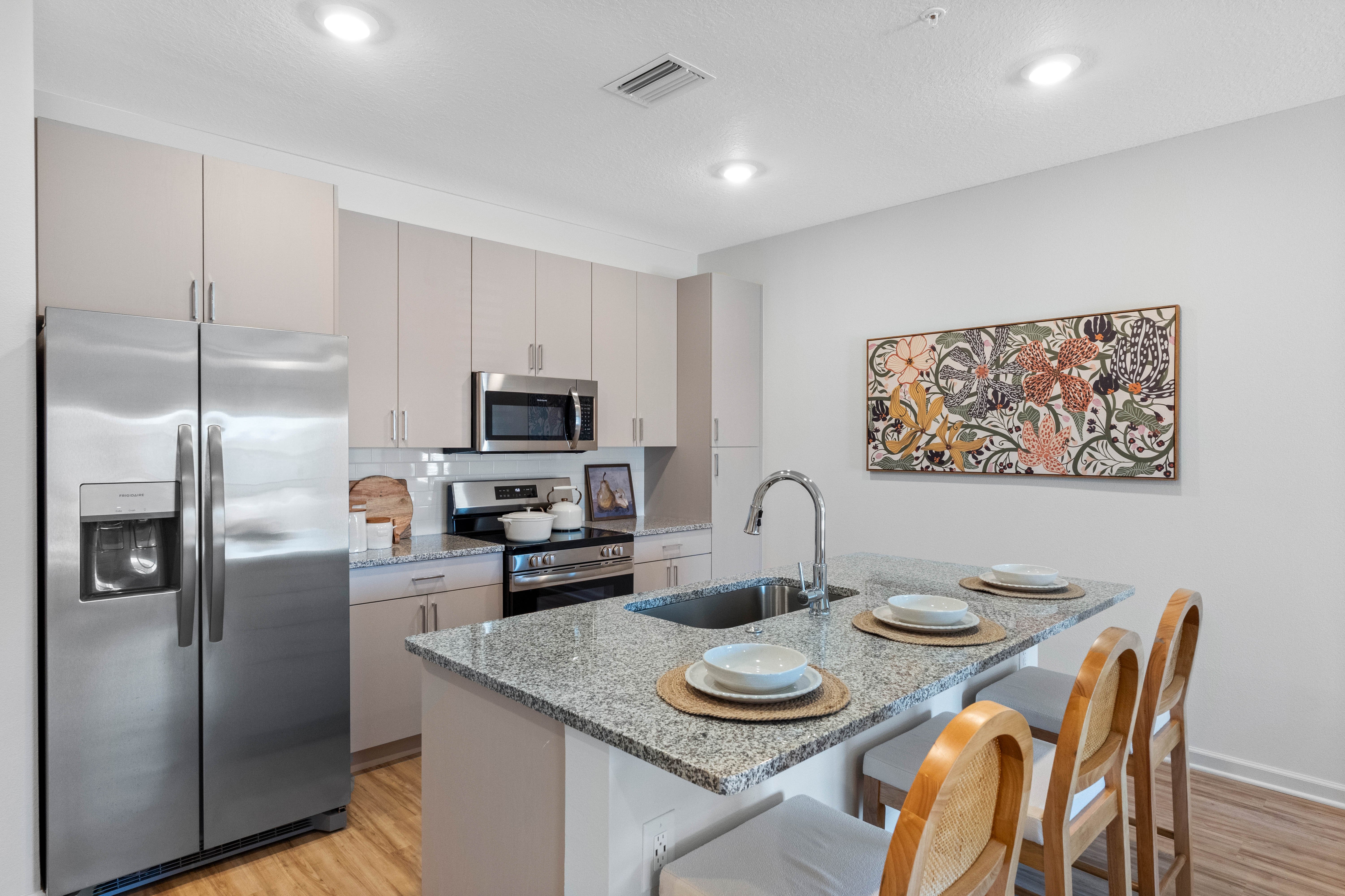 A modern kitchen featuring sleek stainless steel appliances, light colored cabinets, and a granite countertop. A decorative wall art is displayed above the counter with three place settings visible.