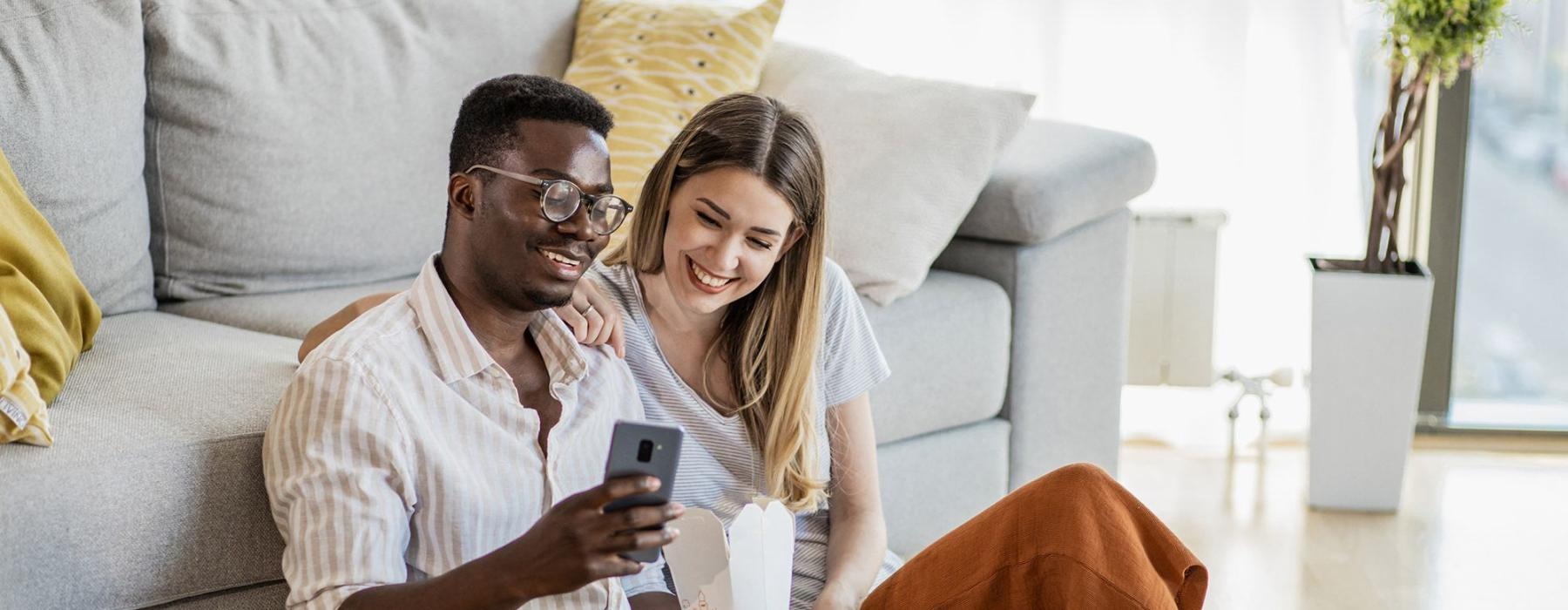 a man and woman with take out, sit against a couch on their living room floor and watch at their cell phone