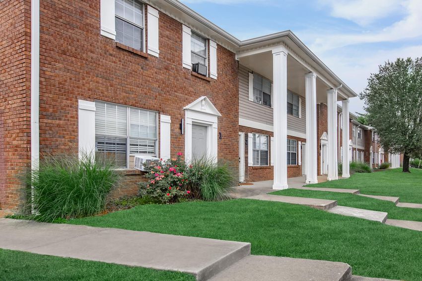 A well-maintained brick apartment building featuring multiple units with white columns and large windows. In front, there are manicured lawns with green grass and decorative rose bushes. The scene is bright and inviting, set against a clear blue sky.