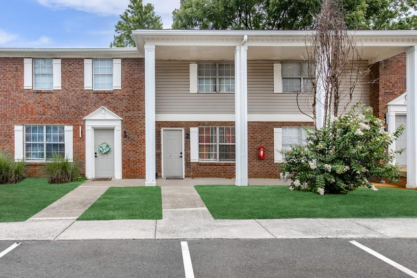 A residential building featuring two stories with brick and siding exterior. The entrance has white columns and a porch area. There are two doors leading into separate units, and the landscaping includes green grass and some shrubs. A fire extinguisher is mounted on the wall.