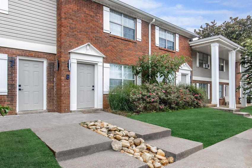 A well-maintained brick apartment building with a manicured lawn. The entrance features two gray doors, surrounded by flowering bushes and decorative rocks along a walkway. The building has white trim and multiple windows, providing a welcoming appearance.