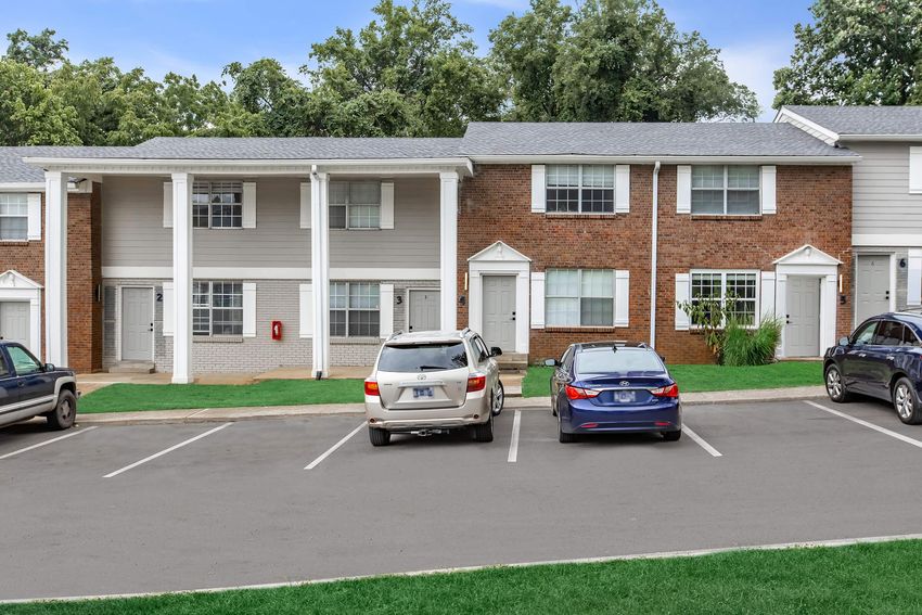 A residential building featuring two-story townhomes with a mix of brick and siding exteriors. Two parked cars, a white SUV and a blue sedan, are visible in front of the units, with greenery lining the walkway. The scene is set in a suburban environment under a clear sky.