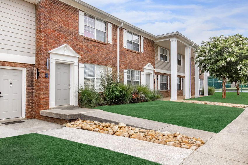 A well-maintained brick apartment building with white columns, featuring several windows and a landscaped yard with green grass and bushes. There are stone pathways leading to the entrance, and the overall setting is clean and inviting. The sky is clear, adding to the bright atmosphere.
