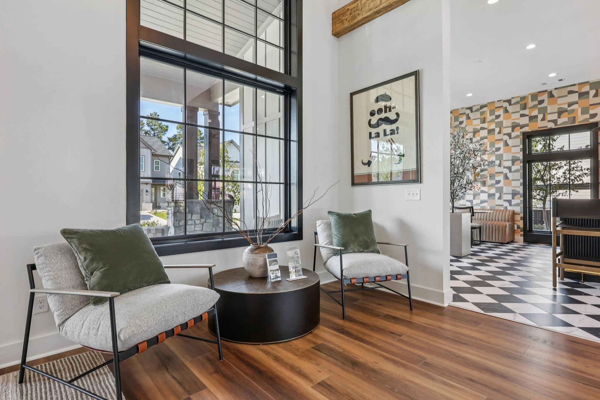 Cozy clubhouse nook featuring two gray armchairs with olive green cushions flanking a round coffee table, a sunny window, and cool geometric wallpaper.