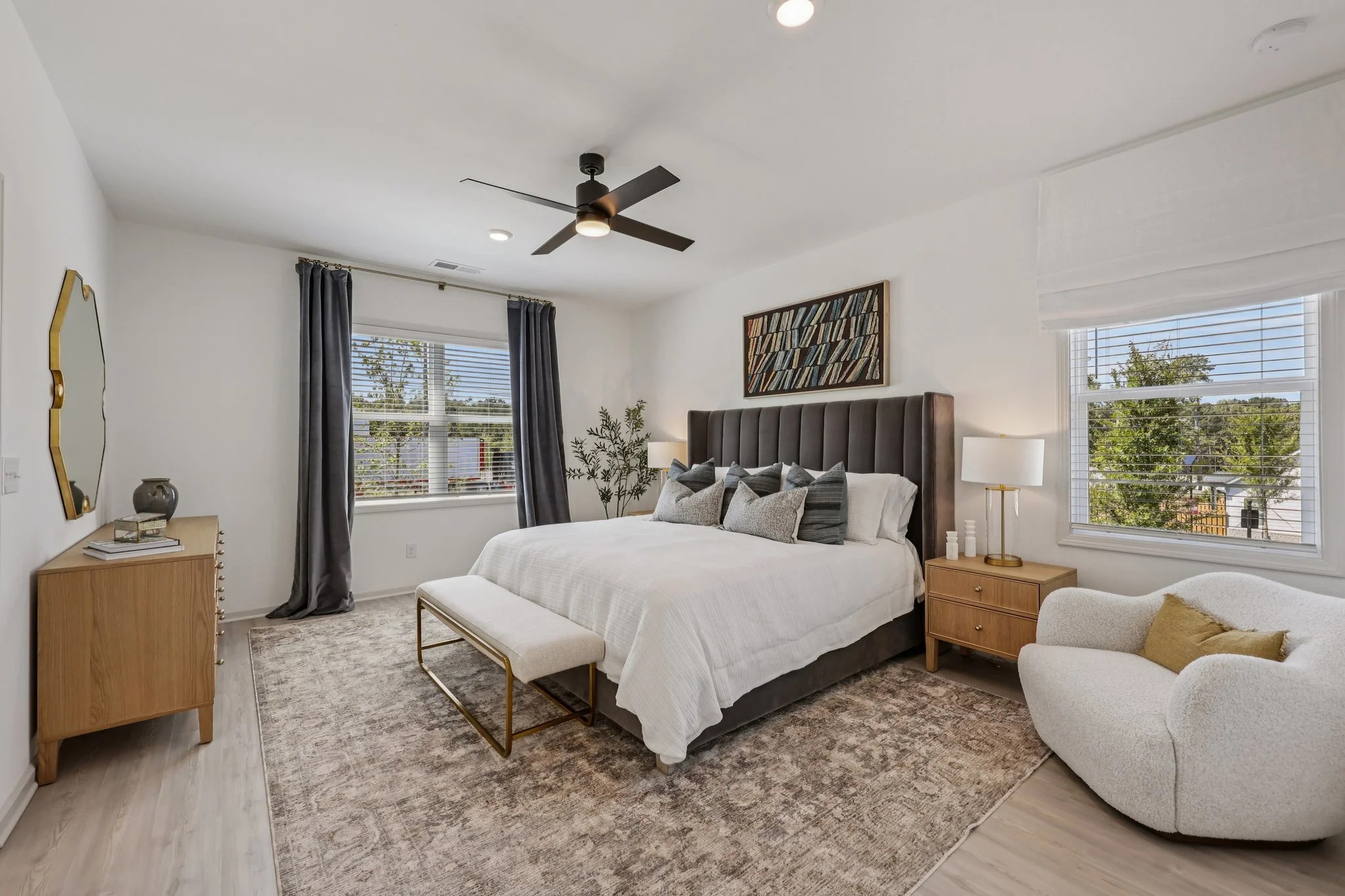 Bright bedroom with mostly white and neutral colors, featuring textural elements like a gray velvet headboard, fluffy white armchair, and patterned rug.