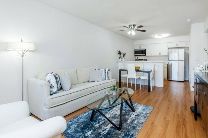 A bright and modern living area featuring a light-colored sofa with blue and white decorative pillows, a glass coffee table on a blue area rug, a dining table with four chairs, and a kitchen area with stainless steel appliances. The walls are white, and there is a ceiling fan and warm wooden flooring.