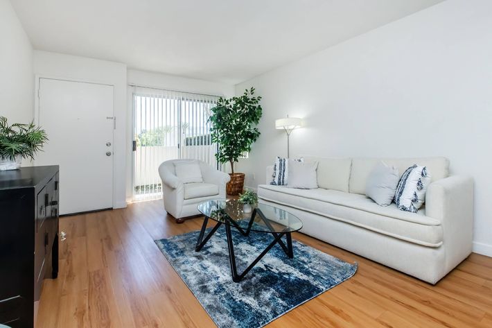 A modern living room with light-colored walls and wooden flooring. It features a white sofa with decorative pillows, a glass coffee table, and a cozy armchair. A tall plant in the corner adds a touch of greenery. Natural light comes in through vertical blinds on a window, enhancing the inviting atmosphere.