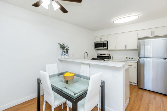 A modern kitchen featuring a dining area with a glass table and white chairs. The kitchen has sleek white cabinets, stainless steel appliances, and a bowl of lemons as a centerpiece. Natural light illuminates the space from a ceiling fan and overhead light fixture. The floor is wooden, enhancing the contemporary feel.