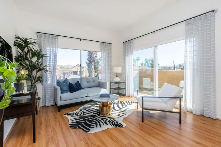 A bright and airy living room featuring a light-colored sofa, a glass coffee table, and tall windows with sheer curtains. Potted plants add a touch of greenery, while light wood flooring complements the modern decor. The view outside includes a sunny landscape, enhancing the inviting atmosphere.