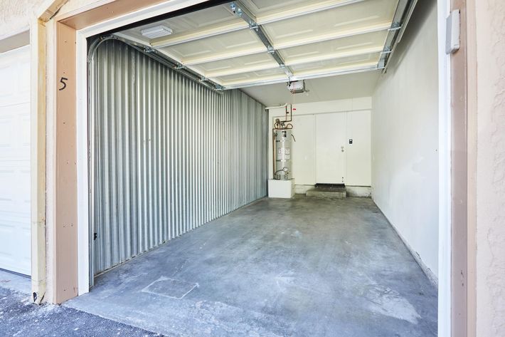 A vacant garage space with a concrete floor, featuring a metal door on one side and a water heater mounted on the wall. The walls are painted white, and the garage door is fully open, revealing the spacious and empty interior.