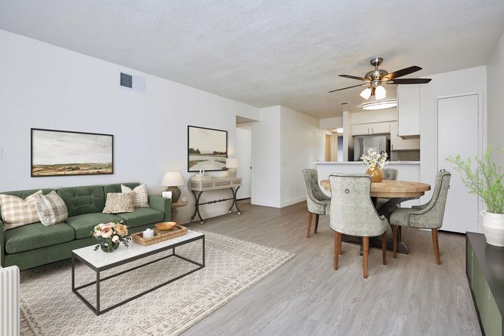 A modern living room and dining area featuring a green sofa with cushions, a coffee table with flowers, a round dining table with gray chairs, and light-colored walls. The space includes framed wall art and a ceiling fan, creating a bright and inviting atmosphere.