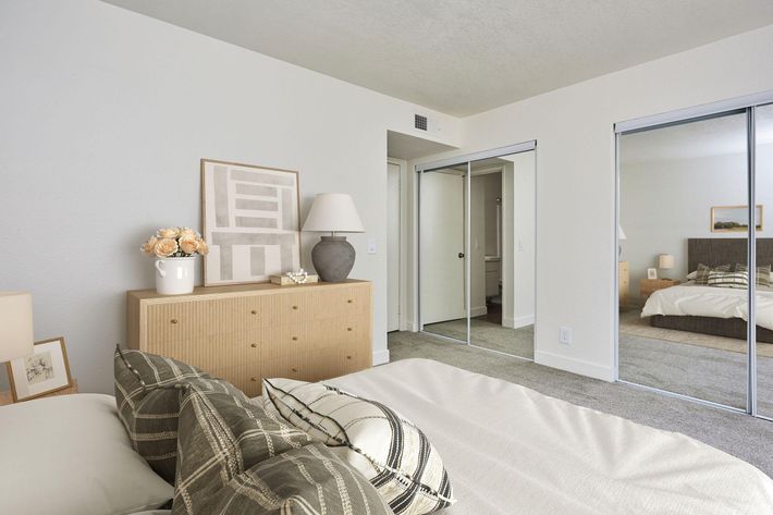 A cozy bedroom featuring a neatly made bed with decorative pillows. There's a light-colored dresser with a lamp and a vase of flowers on top. Mirrored sliding closet doors reflect the room, enhancing the space's brightness. The walls are painted in a soft hue, contributing to a calm atmosphere.