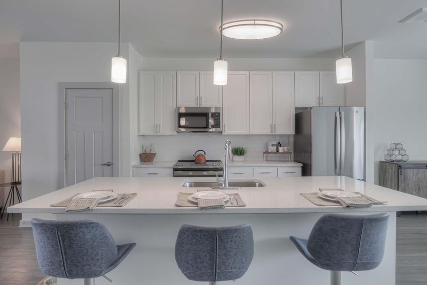 Modern kitchen featuring a large island with three gray bar stools, white cabinets, stainless steel appliances, and pendant lighting. A sink is integrated into the island, and dining plates are set on the counter. Subtle decor includes a plant and kitchen essentials. Soft gray and white tones create a contemporary feel.