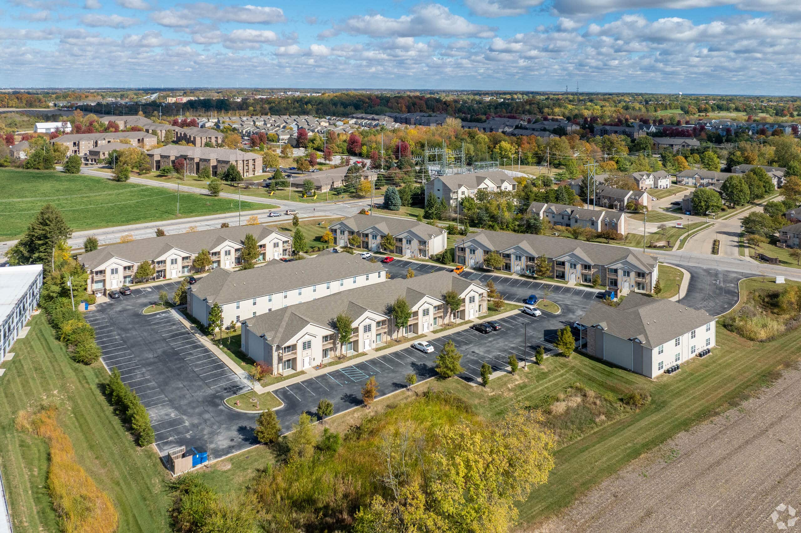 Aerial view of Gallery - McCormick Manor, a residential apartment complex with parking lots, surrounded by trees and open fields under a partly cloudy sky.