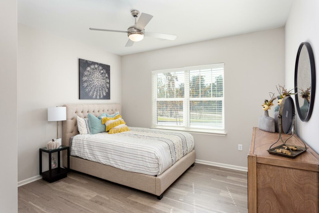 A bright bedroom with a bed, a dresser, a ceiling fan, hardwood-style flooring, and a window.