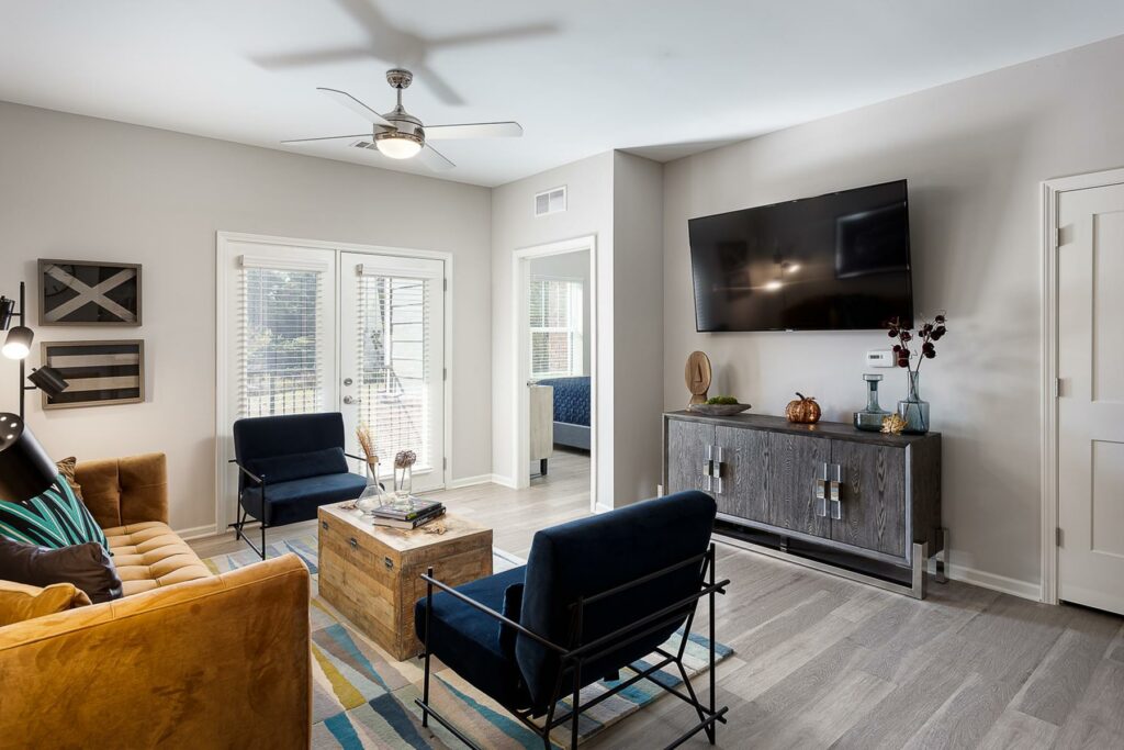 A spacious living room with a couch, two chairs, a table, a TV, and a large window, on hardwood-style flooring.