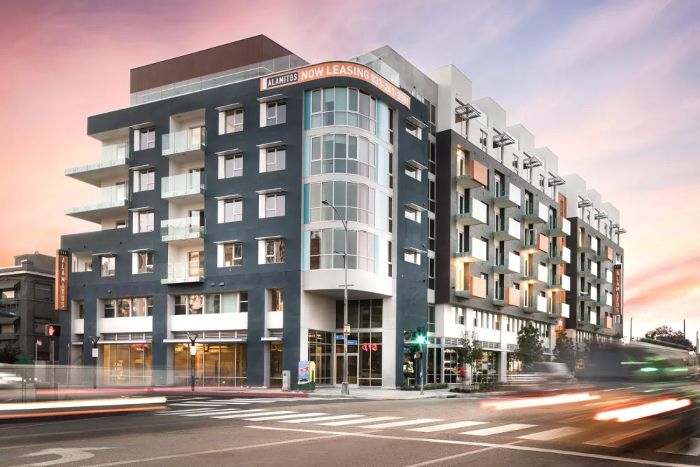 Street view of The Alamitos Apartments, a contemporary mid-rise community with private balconies, large windows, and a prominent "Now Leasing" sign on the corner building.  Photo 3 – Resort-S