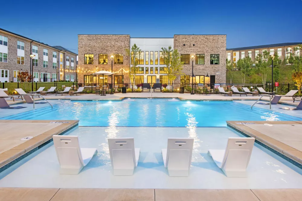Sparkling resort-style swimming pool at The Statesman Apartments with in-water lounge chairs, surrounding sun deck, and string lights creating a relaxing evening atmosphere.