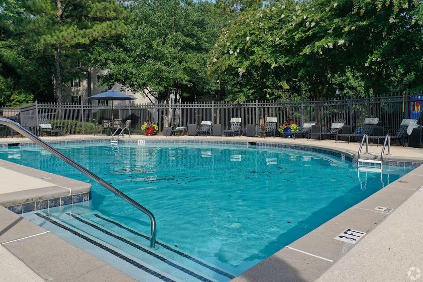A clear blue swimming pool surrounded by a concrete deck, with steps leading into the water. There are several lounge chairs arranged around the pool and a fence in the background. Trees provide shade, and there are some potted plants nearby. No people are present in the scene.