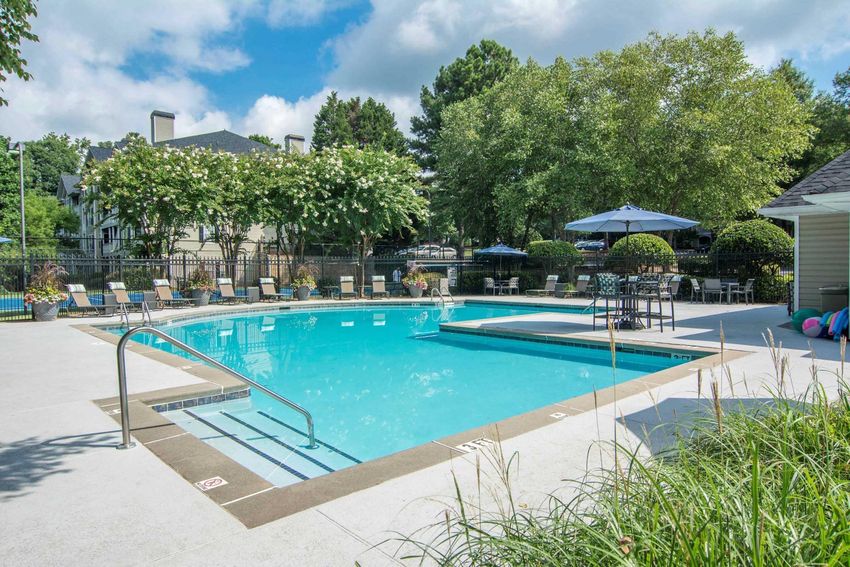 A bright outdoor swimming pool surrounded by lush greenery and lounge chairs. Umbrellas provide shade around the pool area, which is fenced for safety. In the background, there are residential buildings and colorful flowers. The sky is partly cloudy, creating a serene atmosphere.