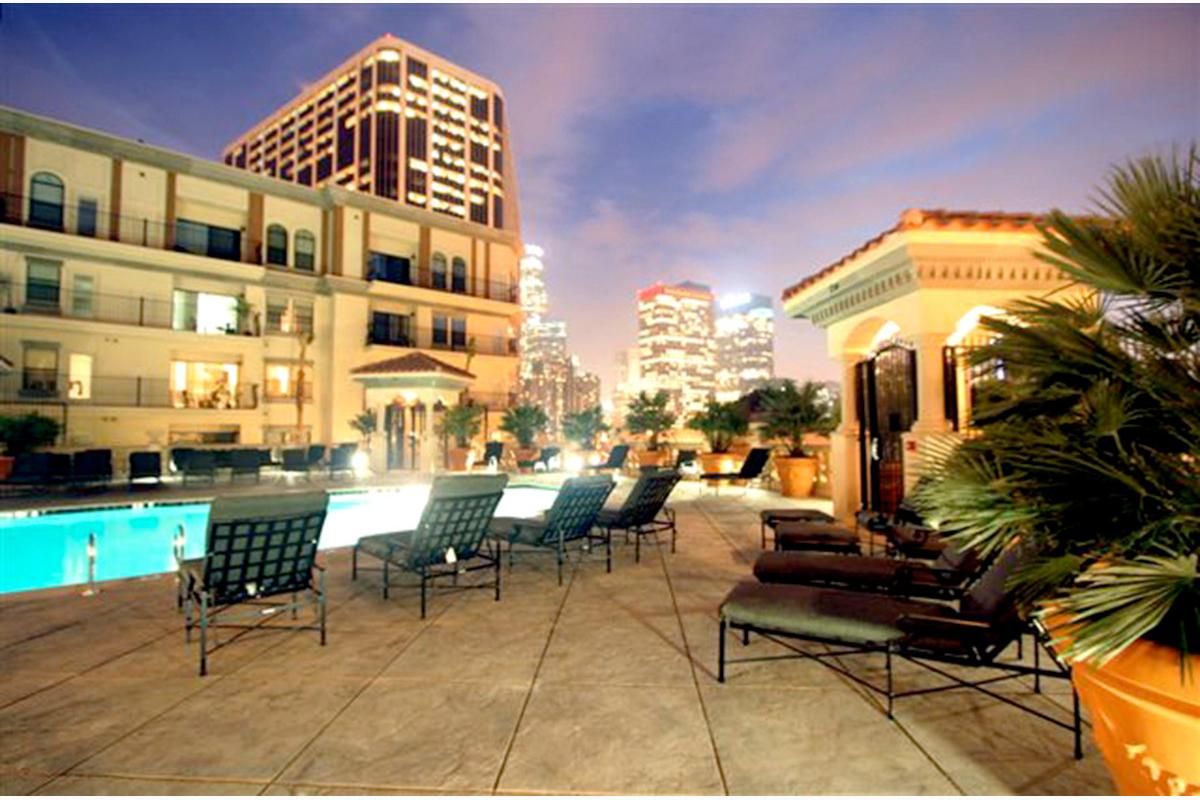 A serene rooftop pool area at dusk, featuring loungers arranged around a sparkling pool. Skyscrapers and city lights are visible in the background, creating a vibrant urban atmosphere. Lush greenery and decorative architecture enhance the relaxing ambiance of the space.
