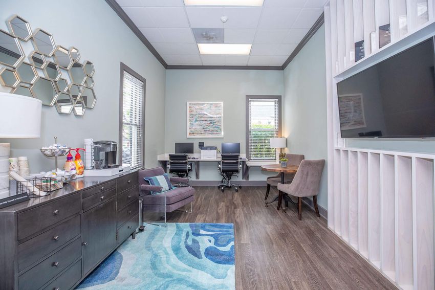 A modern office space featuring light green walls, a patterned rug, a dark wooden dresser with refreshments, two computer desks, and a cozy chair. The room has large windows allowing natural light, and decorative hexagonal mirrors on one wall.