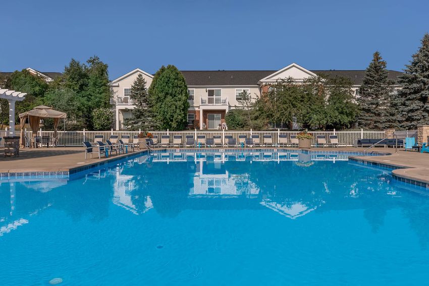 A clear swimming pool surrounded by lounge chairs, with residential buildings and greenery in the background under a bright blue sky.
