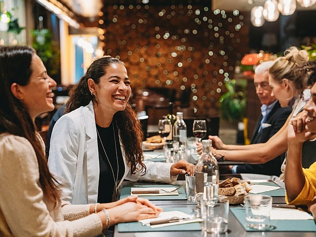 Group of cheerful friends enjoying a dinner party in a stylish, warmly-lit restaurant with elegant decor and wine glasses.