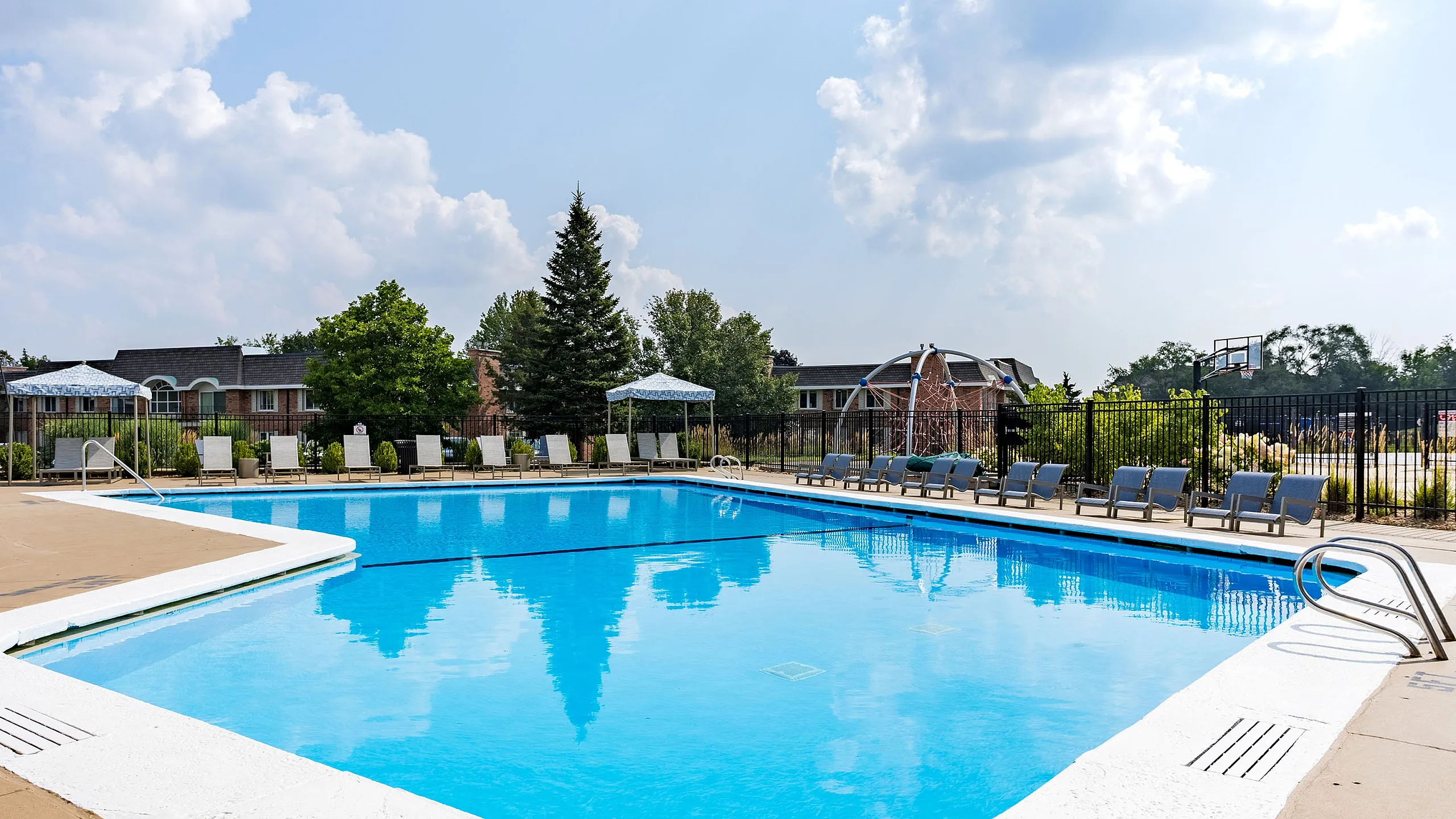 Inviting outdoor swimming pool area with sun loungers, surrounded by lush greenery and bright skies.