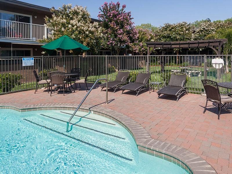 Outdoor apartment pool area with lounge chairs, brick-paved deck, and a fence.