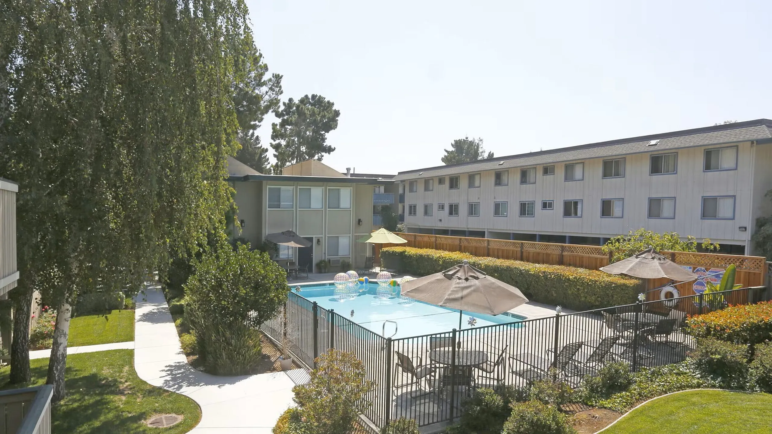 Apartment complex with a fenced swimming pool, surrounded by sunbeds and umbrellas. Pathways lead through well-maintained green gardens. Tranquil ambiance.
