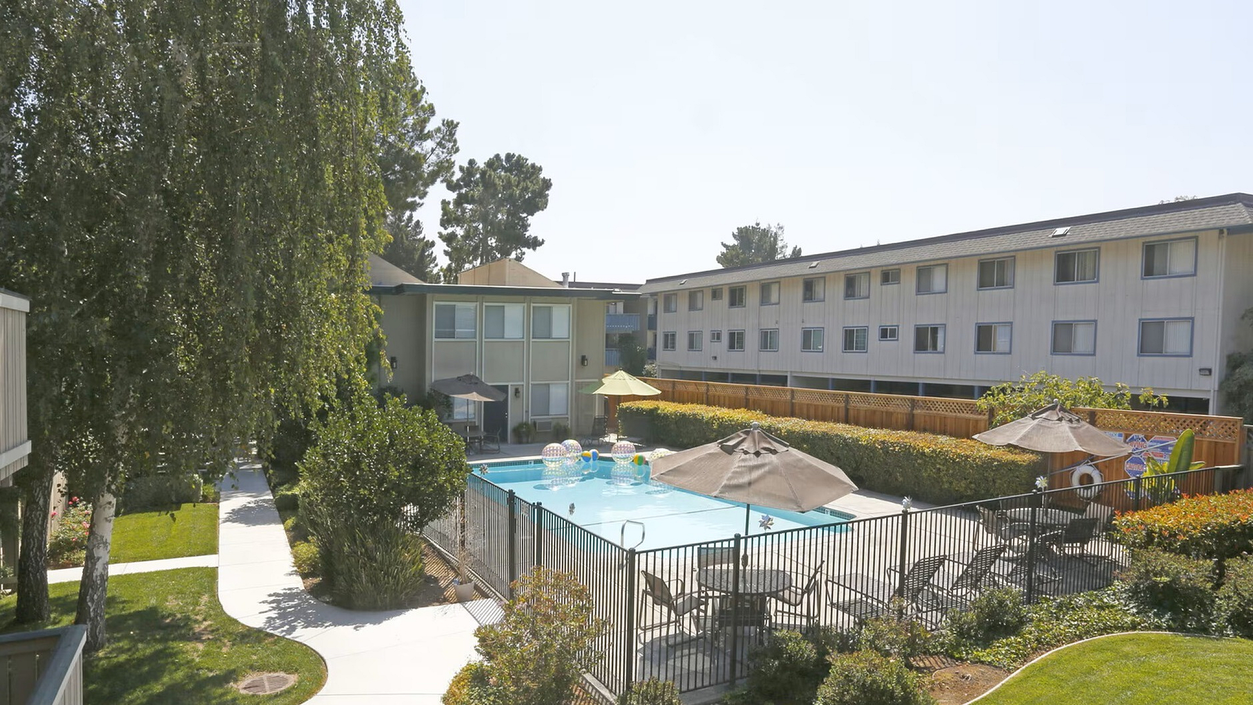 Apartment complex with a fenced swimming pool, surrounded by sunbeds and umbrellas. Pathways lead through well-maintained green gardens. Tranquil ambiance.
