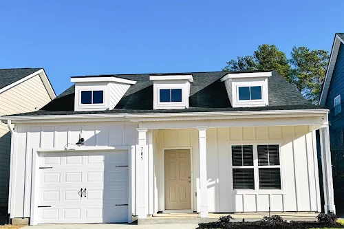 A light color house with a garage, dormer windows, and a front door under a clear blue sky.
