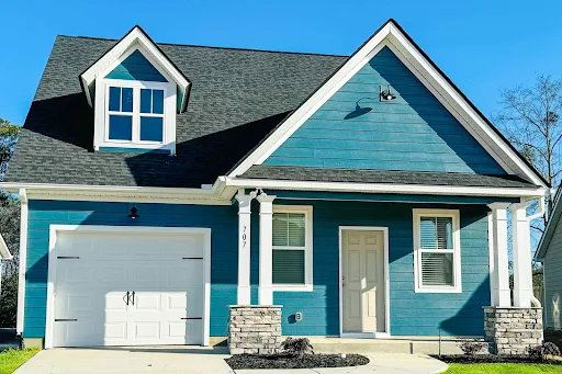 Blue apartment complex exterior featuring a garage, dormer window, and landscaped entrance under clear skies.