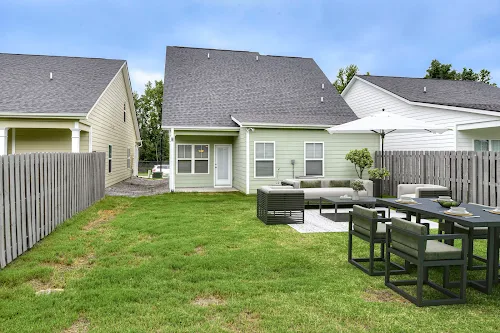 A well-maintained backyard featuring a seating area, dining table, and a patio umbrella behind a green house.