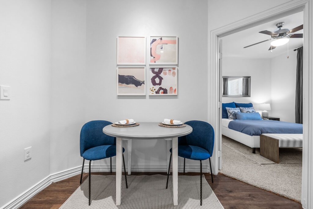 Round dining table with two blue chairs. View into bedroom.