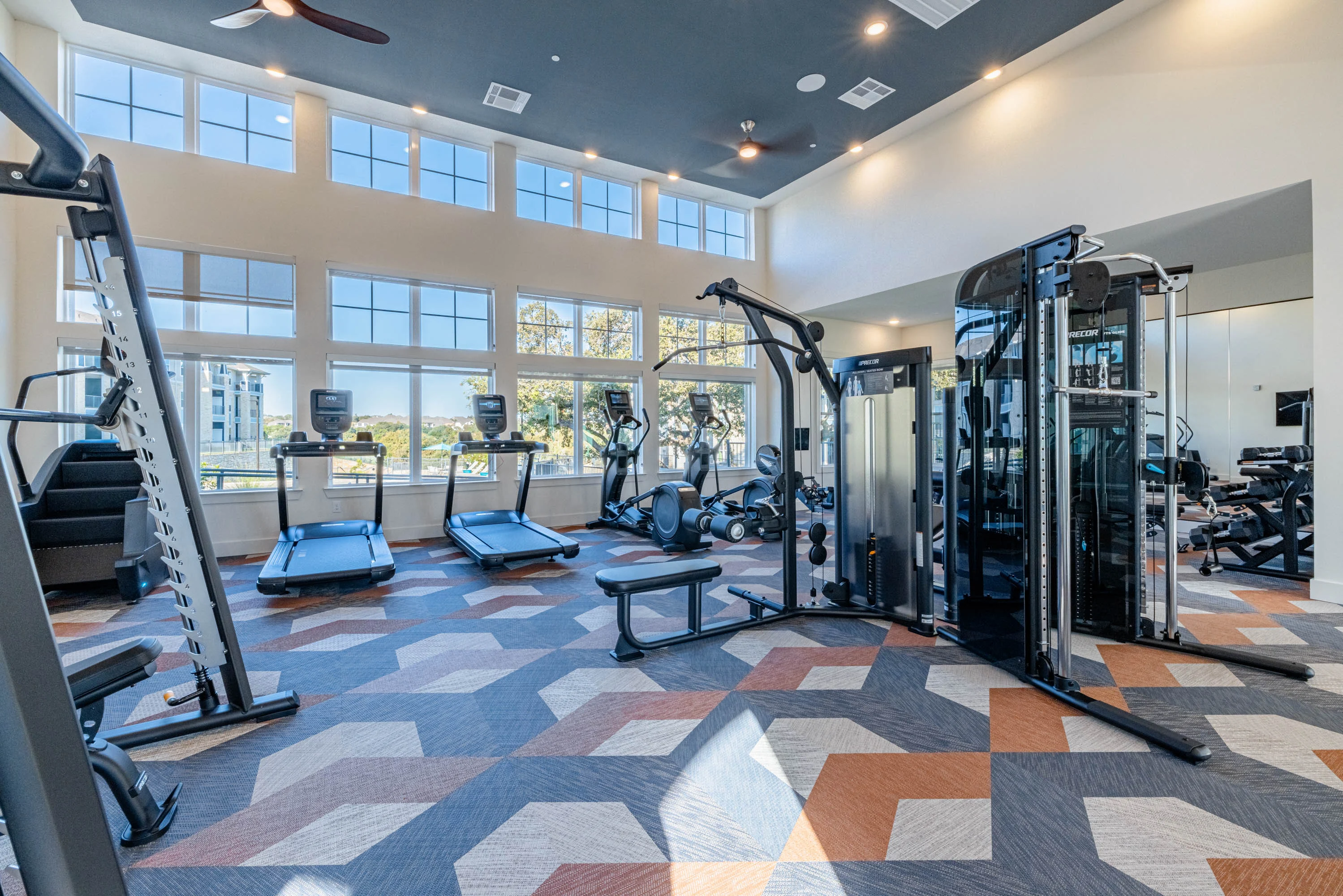 Apartment complex gym with treadmills, weights, and large windows offering ample natural light.