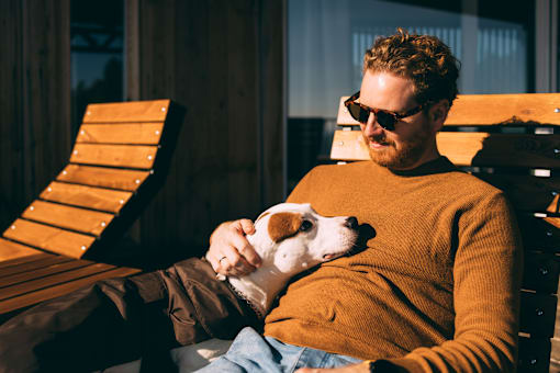 A man in a brown sweater is sitting on a wooden chair holding a small dog.