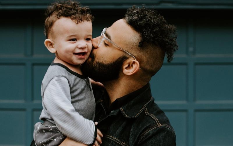 A man with a beard and glasses holds a smiling toddler in his arms, kissing the child's cheek.