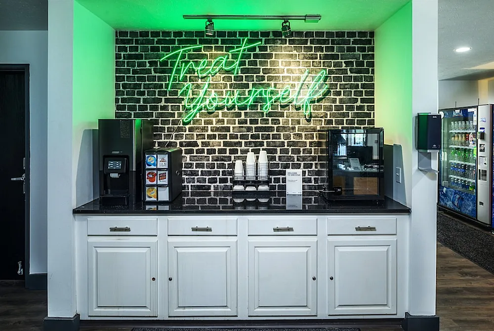 Modern refreshment station with sleek cupboards beneath a vibrant 'Treat Yourself' neon sign, featuring coffee and beverage options in a stylish lounge setting.