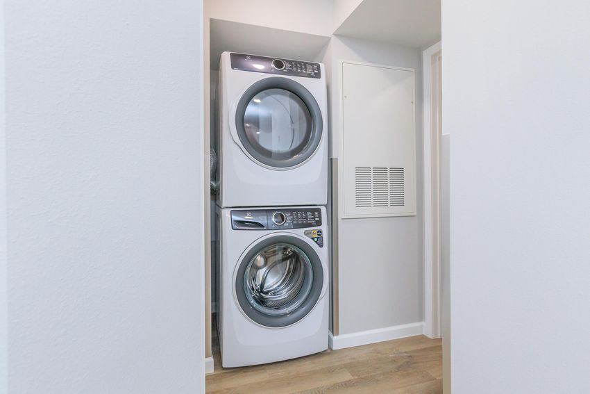 A stackable washer and dryer unit located in a narrow laundry space with light-colored walls and wooden flooring.